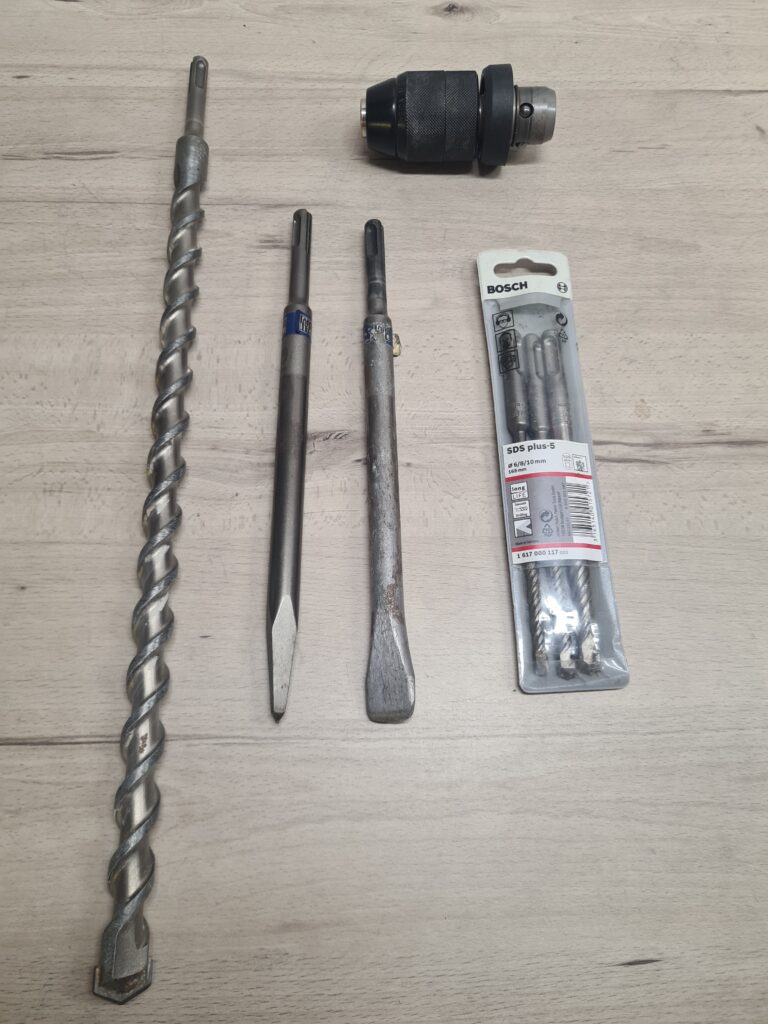 Verschiedene SDS-Plus Bohrer, Spitz- und Flachmeißel sowie ein Wechselbohrfutter auf einer Holzfläche.